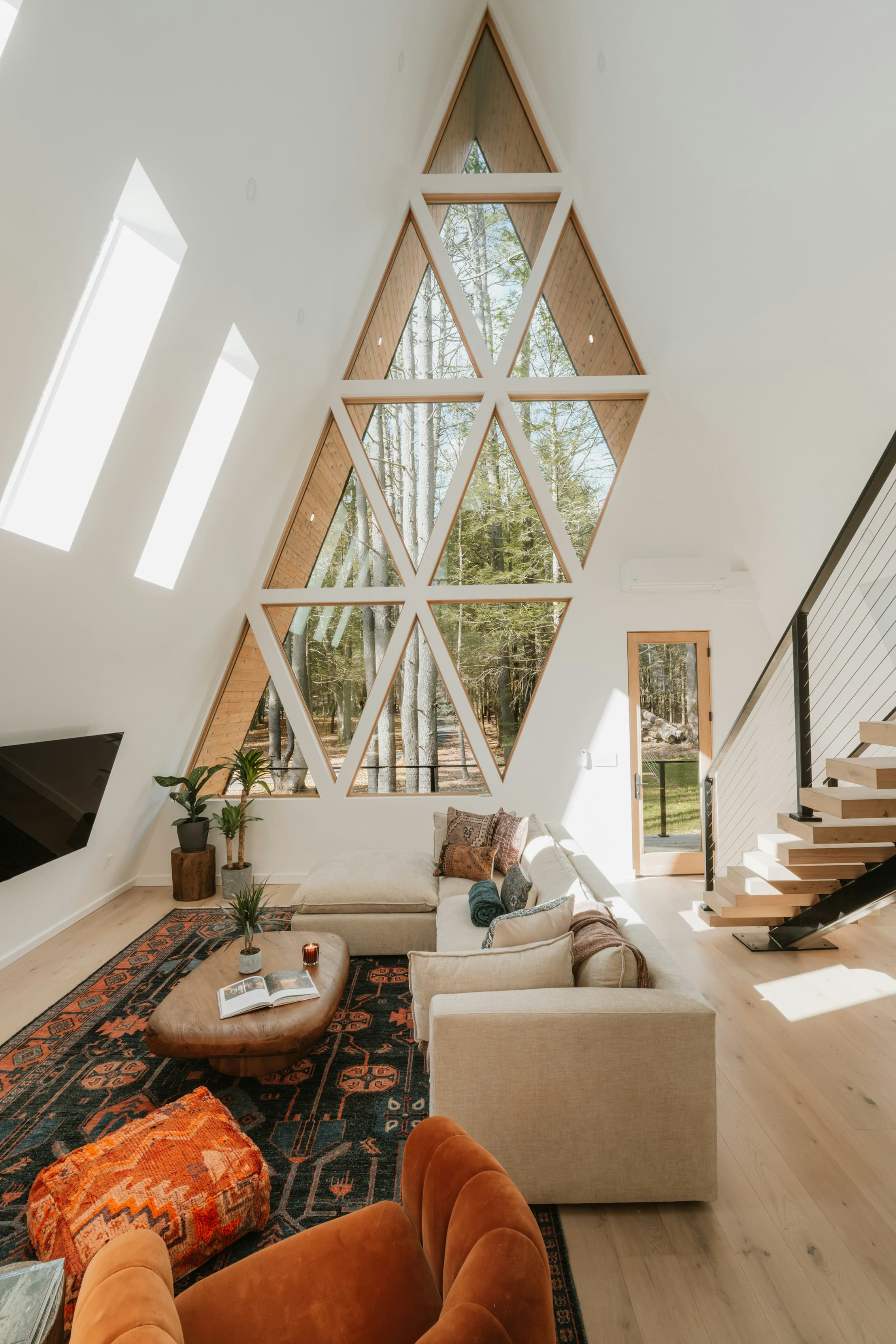 A-frame vacation rental living room with floor-to-ceiling geometric windows, forest view, and layered boho interior